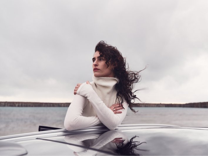 Image of a woman outdoors leaning on a car roof with wind blowing in her hair. The sea is in the background.