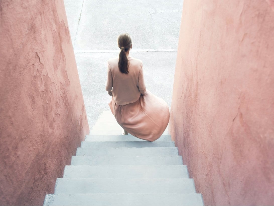 image of a woman walking down stairs in a concept location. She is dressed in pink, the walls are pink stone and the staircase is in off white stone.