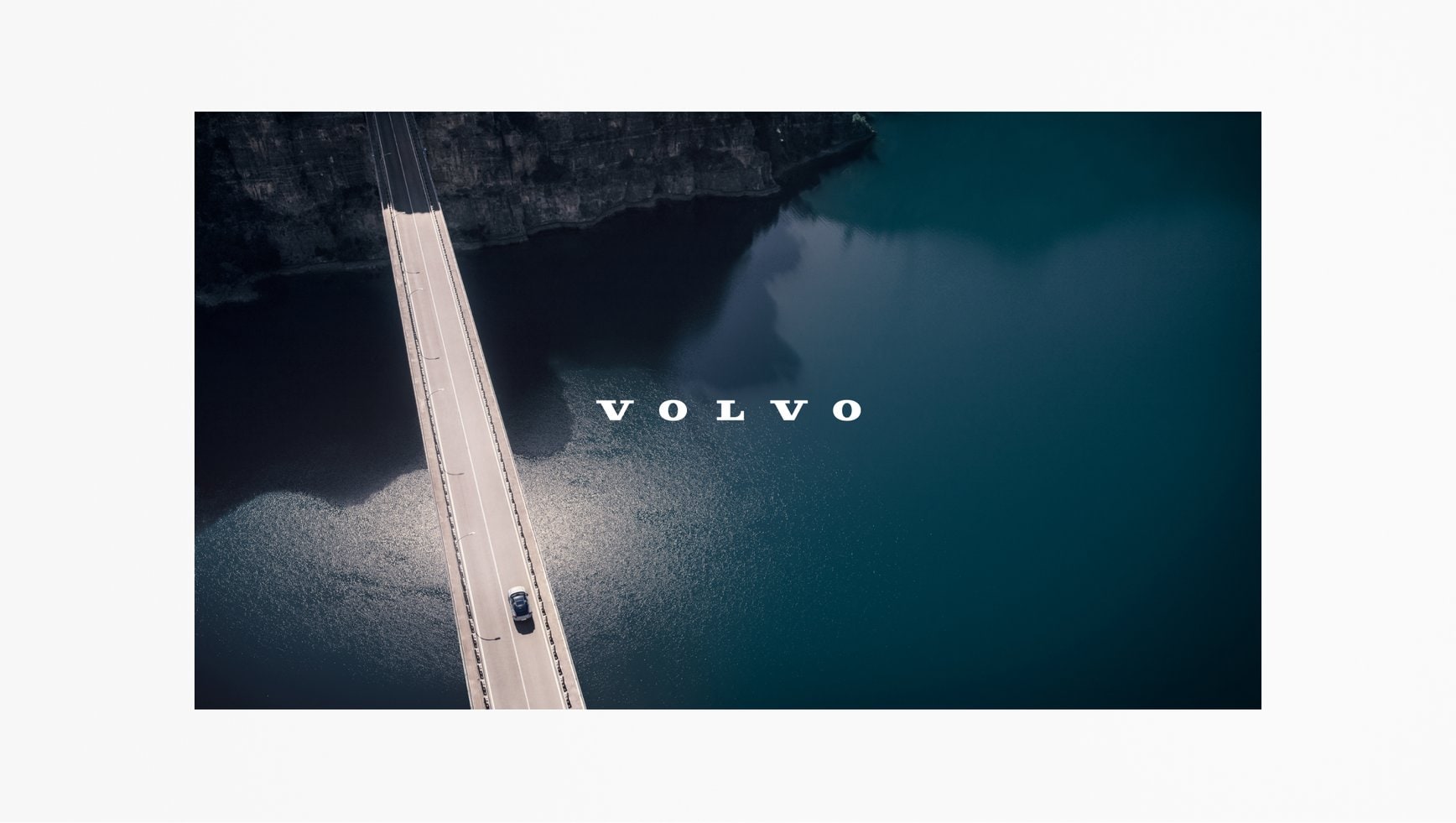 This aerial image represents an end of video context (in a widescreen format). The photo is of a car driving over a suspension bridge, with a body of water underneath. The Volvo Spread Word Mark is positioned in the centre of the photo. It contrasts well, being set in white, on the dark water in the background.